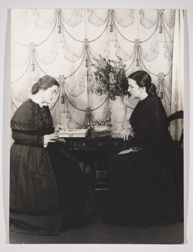 Two women in vintage clothing are seated opposite each other with an ornate floral curtain backdrop. The woman on the left is looking down at a book she is holding, while the woman on the right gazes directly at her. A vase with flowers sits between them on a small table, and the setting suggests a scene from the late 19th or early 20th century.