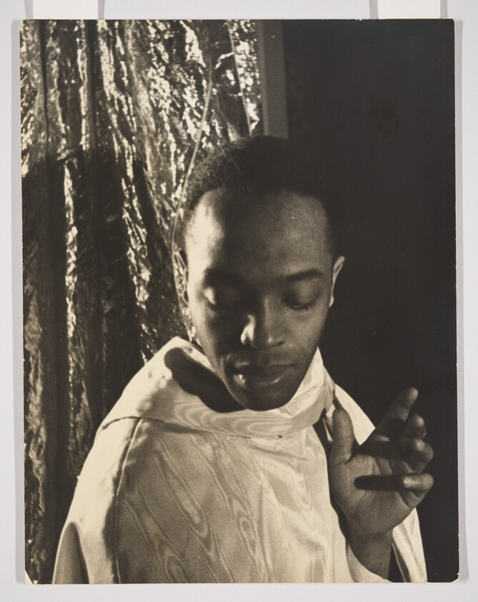 A vintage black and white photograph of a person with a contemplative expression, looking downward with their head slightly tilted. They are wearing a light-colored, textured garment with a high collar, and their right hand is raised near their face with two fingers extended. The background features a dark, textured curtain to the left, contrasting with the lighter, smooth surface to the right.