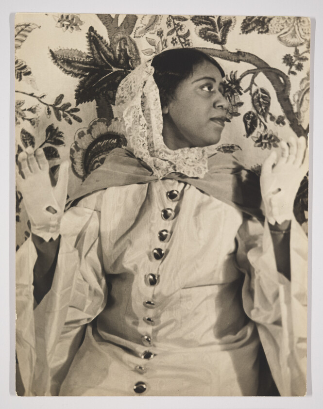 A vintage black and white photograph of a woman wearing a bonnet with lace trim and a dress with button details. She is posed against a patterned backdrop, looking to the side with a thoughtful expression, and her hands are raised slightly in front of her.