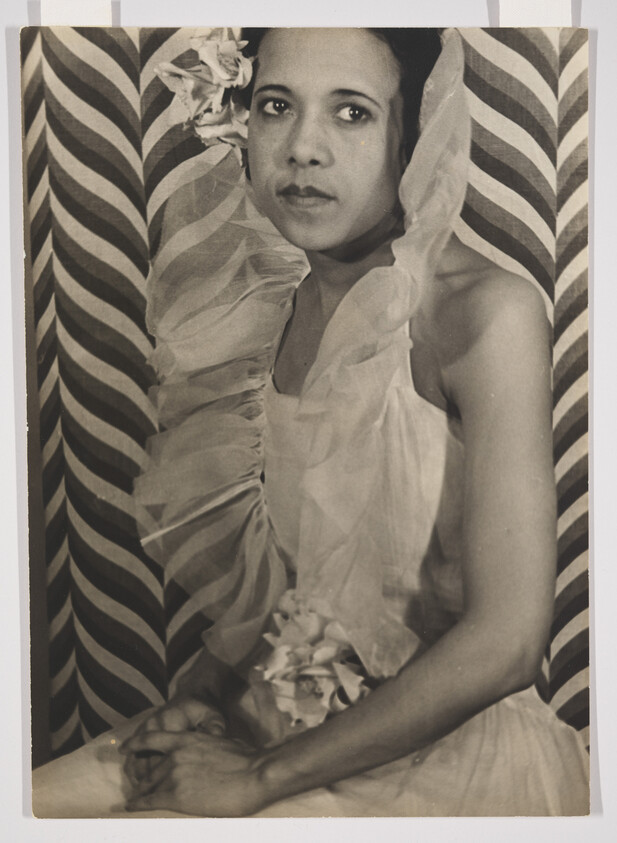 A vintage black and white photograph of a woman with a contemplative expression, wearing a sheer fabric with floral embellishments, seated in front of a striped backdrop.