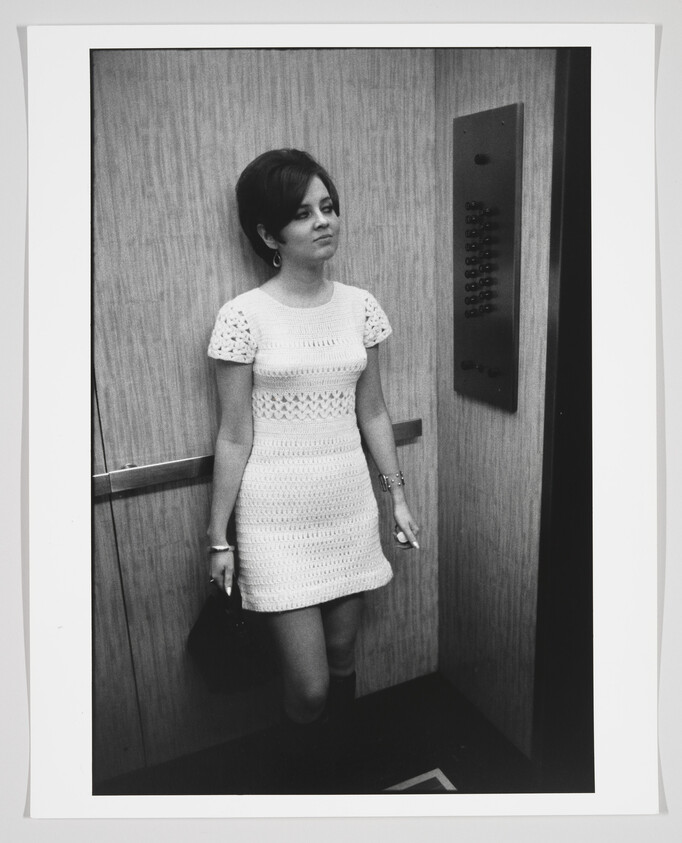 A black and white photo of a woman standing inside an elevator, looking to her side with a slight smile. She is wearing a short-sleeved, knee-length white dress with a patterned design and is holding a purse in her left hand. A panel of elevator buttons is visible to her right.