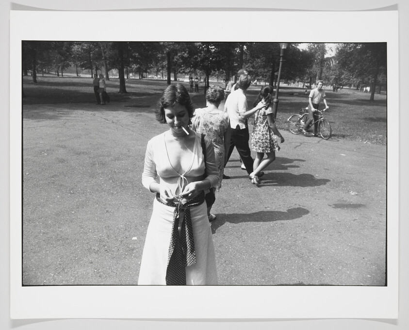 A black and white photograph capturing a moment in a park with a woman in the foreground tying a sweater around her waist. She is looking down and has a cigarette in her mouth. In the background, various other people can be seen walking and enjoying the park, including a couple holding hands and a person with a bicycle. Trees and open grass areas suggest a leisurely, sunny day.