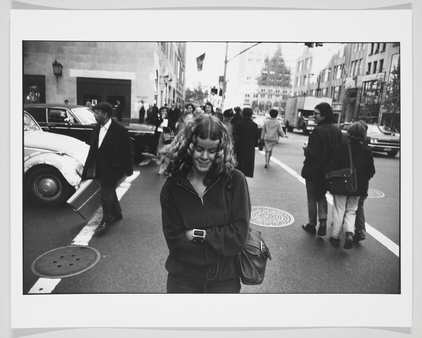 Black and white photo of a smiling young woman walking on a city street, looking down at a device in her hands. She is wearing a dark jacket and has curly hair. In the background, various pedestrians are crossing the street, and there's a classic car with its door open. City buildings and a traffic light are visible in the distance.