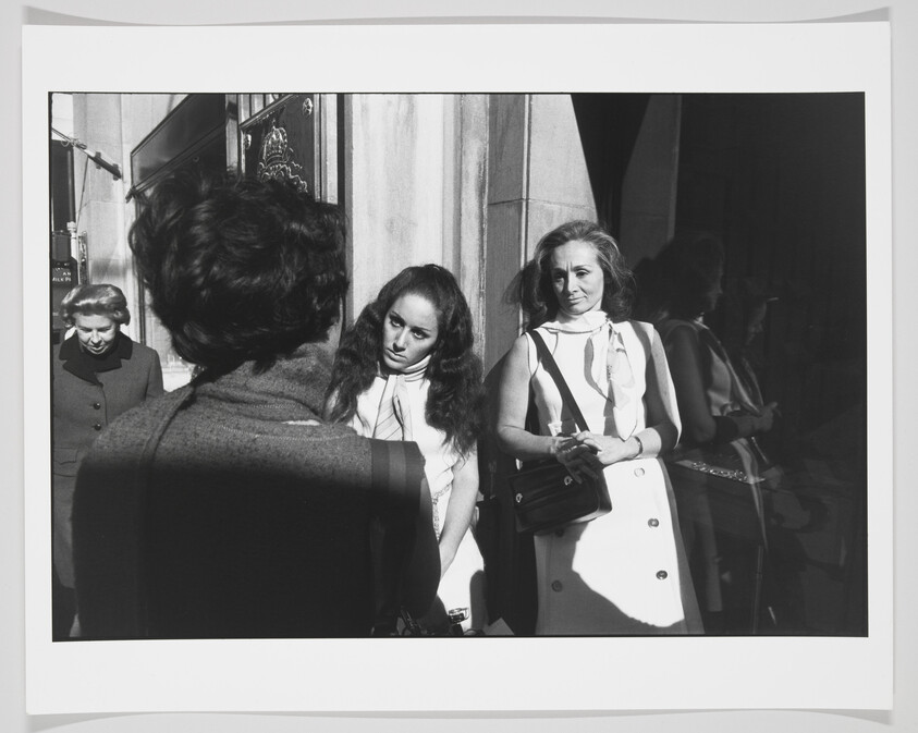 A black and white photograph capturing a moment on a sunny street where two women are standing and looking towards a person with their back to the camera, possibly engaged in a conversation. The women are dressed in light-colored, stylish clothing indicative of mid-20th-century fashion, with one woman holding a purse. The scene is marked by strong contrasts of light and shadow, highlighting the expressions on the women's faces and the architectural details of the building behind them.