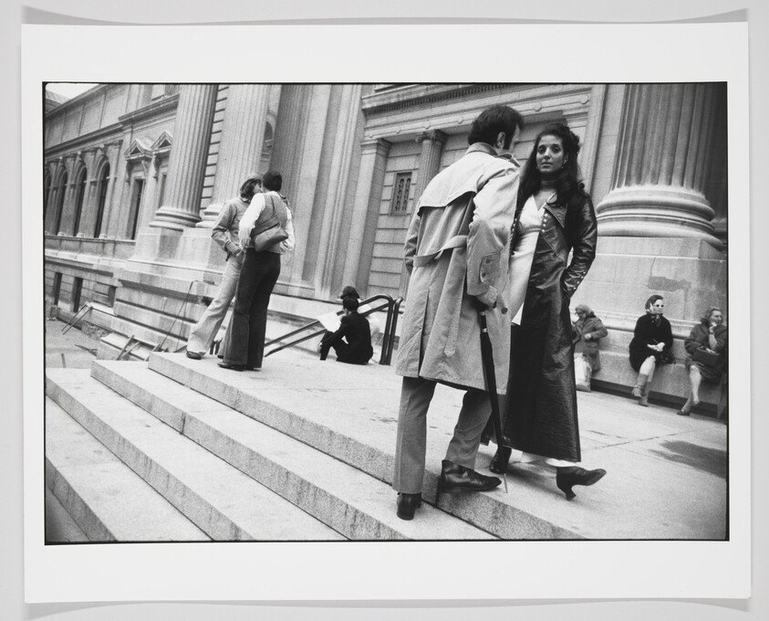 A black and white photograph capturing a candid street scene with various people on the steps of a building with classical architecture. In the foreground, a couple stands close together, the man facing away from the camera and the woman looking directly at the lens with a neutral expression. Other individuals are seated or standing in the background, seemingly engaged in their own activities.