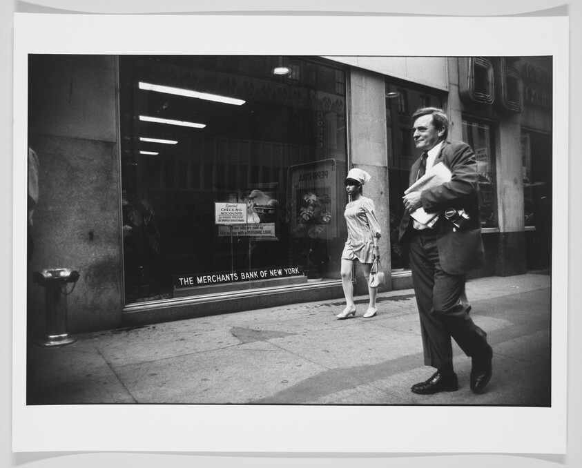 Black and white photograph capturing a moment on a city street with a man in a suit walking briskly past a storefront window of The Merchant's Bank of New York, while a woman in light clothing stands by the window, looking in another direction.