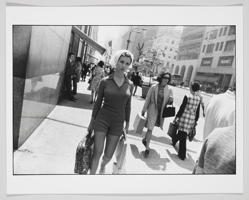 A black and white photograph capturing a bustling city street scene with a woman in the foreground looking directly at the camera. She is wearing a fitted, long-sleeve top and shorts, carrying a large patterned bag in her right hand. The background is filled with various pedestrians, some in the shade of buildings and others in sunlight, suggesting a busy urban environment during daytime.