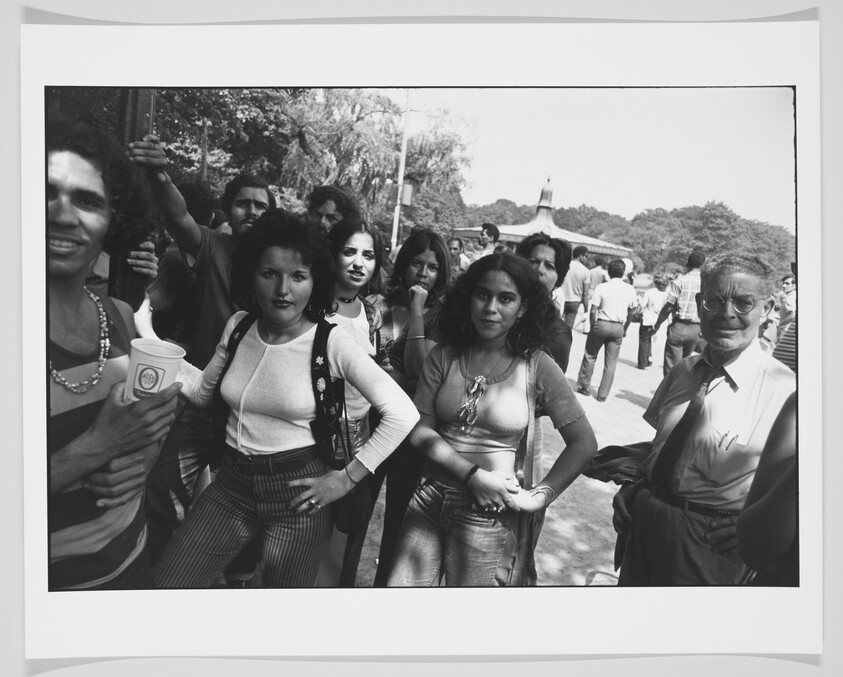 A black and white photograph capturing a diverse group of people, possibly at a public event or gathering. The individuals are of various ages and ethnicities, with some looking directly at the camera. The foreground features a man on the left holding up a cup and a woman in the center with her hands on her hips. In the background, more people can be seen mingling, with trees and a structure with a flagpole in the distance. The expressions range from smiles to neutral gazes, suggesting a casual and relaxed atmosphere.