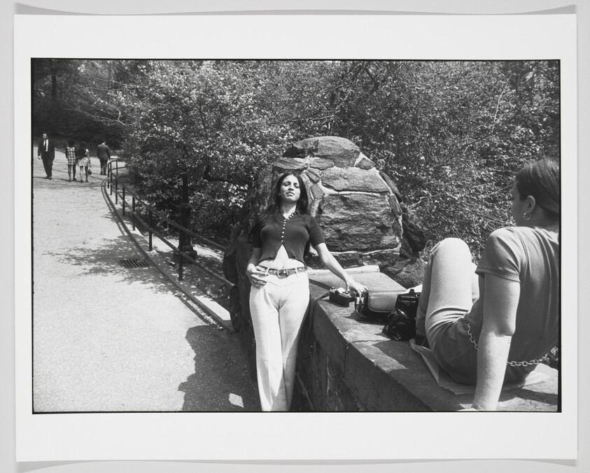 A black and white photo depicting a woman leaning against a stone wall in a park-like setting, with another person sitting on the wall facing away from the camera. The woman leaning is in the foreground, looking to the side with a relaxed posture, while the sitting person's legs are in the foreground. In the background, there are people walking along a path lined with trees and a metal railing.