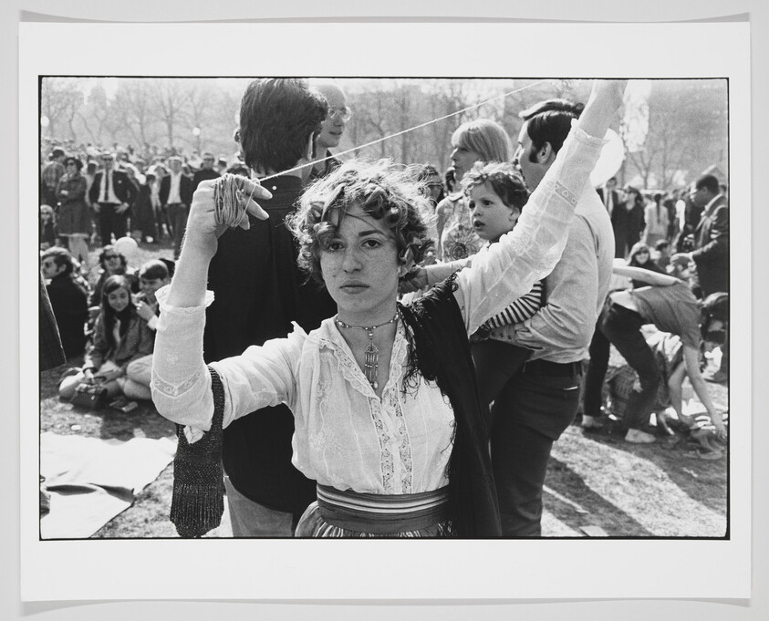 A black and white photograph capturing a moment at an outdoor gathering, possibly a protest or festival. In the foreground, a young woman with curly hair, wearing a white blouse and a dark vest, looks directly at the camera with a serious expression. Her right hand is raised above her head, holding onto a string or thin rope that extends out of frame. Behind her, various people can be seen in the background, some standing and others sitting on the ground, suggesting a casual and possibly communal atmosphere. The attire and hairstyles suggest the image might be from the 1960s or 1970s.