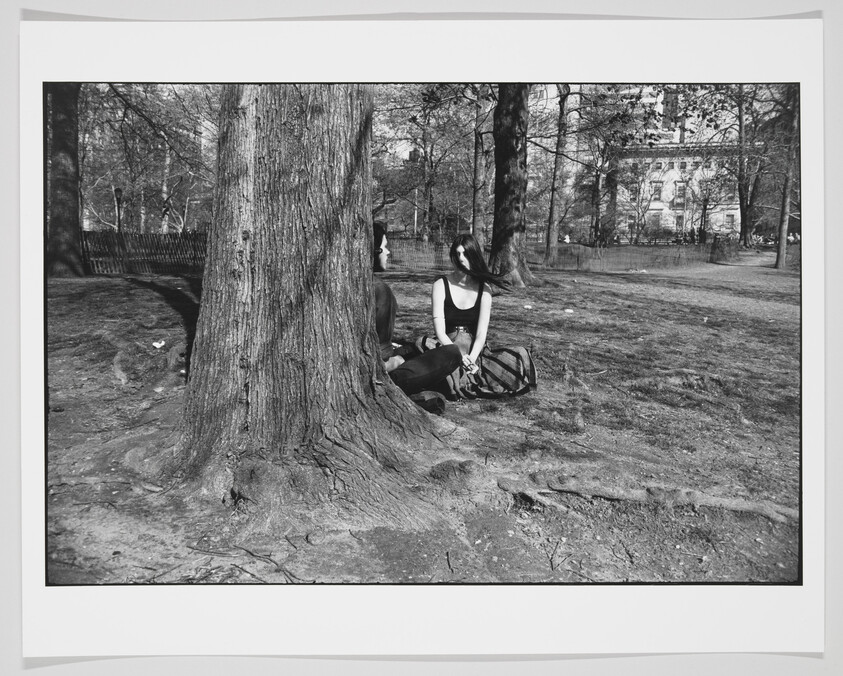 A black and white photo of a person sitting at the base of a large tree in a park, with their hair blowing across their face and a backpack beside them. The background shows a fence and leafless trees, suggesting it might be late fall or winter.