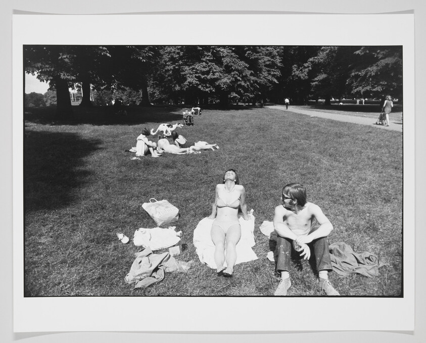 A black and white photo of people relaxing on the grass in a park on a sunny day. Two individuals are prominently featured in the foreground, one sitting upright and the other reclining, with others scattered in the background under the shade of trees. There are various personal items strewn about on the grass, suggesting a leisurely day out.