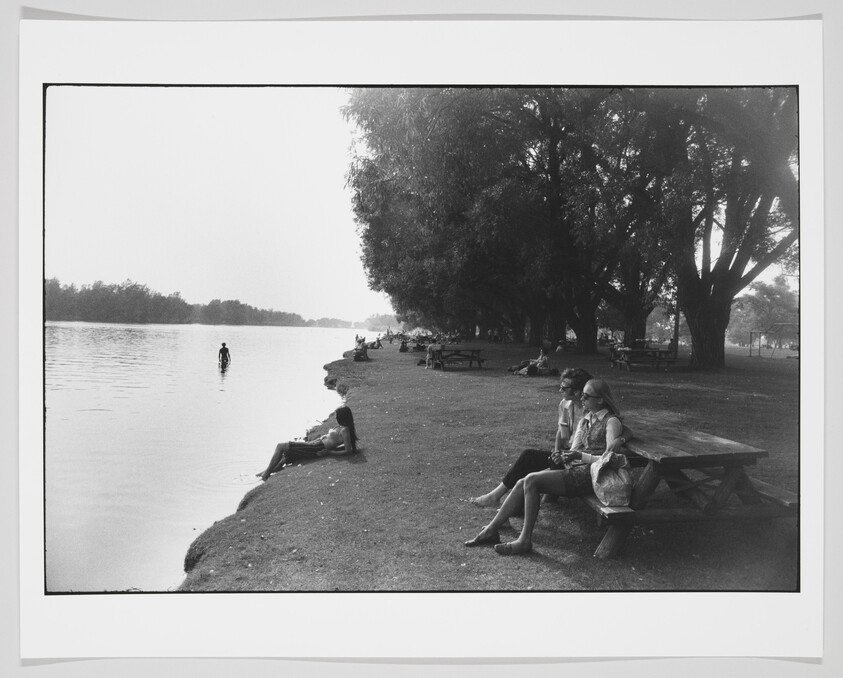 Black and white photograph of people relaxing by a calm river. On the left, a person lounges on the grassy bank, while another stands in the water. To the right, two individuals sit on a picnic bench under the shade of large trees. The park atmosphere is serene with open space and scattered visitors enjoying the outdoors.