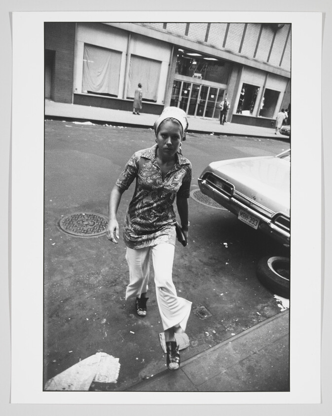 A black and white photograph capturing a woman walking on a city street, looking directly at the camera with a neutral expression. She is wearing a patterned blouse, white pants, and sandals, with a headscarf tied around her hair. In the background, there's a classic car parked on the side of the road, and a few pedestrians are visible in the distance near a storefront with covered windows. The urban setting suggests a scene from a past decade.