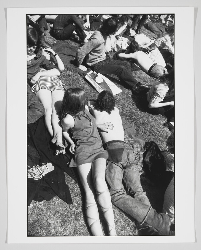 A black and white photograph of a group of people lounging on the grass, with some sitting and others lying down. In the foreground, a person in a sleeveless dress and knee-high socks is seated on the grass leaning against another individual, while various personal items like shoes and a bag are scattered around. The atmosphere appears relaxed and casual, indicative of a sunny day in a park or outdoor gathering.