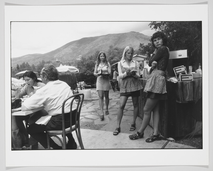 A black and white photograph capturing a candid moment at an outdoor event with mountains in the background. On the left, a man and a woman are seated at a table, seemingly engaged in conversation. In the center, a woman walks towards the camera holding a tray, while another woman stands in the foreground looking at a device in her hands. On the right, a woman leans against a wooden barrel, arms crossed, looking directly at the camera. There are umbrellas and stalls in the distance, suggesting a fair or market atmosphere.