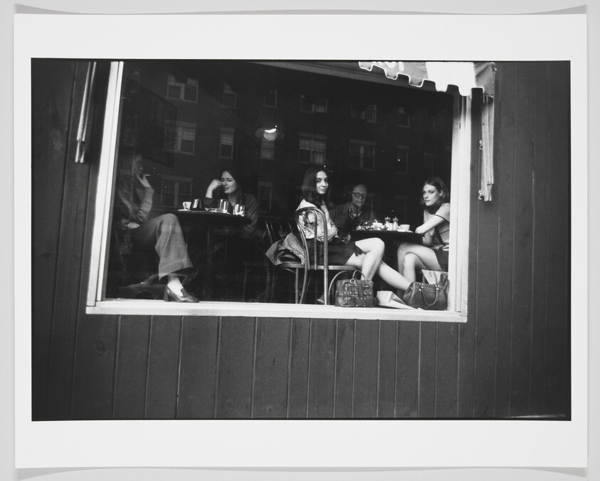 A black and white photograph capturing four individuals seated inside a cafe, viewed through a large window from the outside. Two are facing the window directly, while the other two are in profile, engaged in conversation or looking away. They are surrounded by coffee cups and small plates on the tables, with reflections of buildings visible in the window's glass. The scene conveys a casual, relaxed atmosphere.