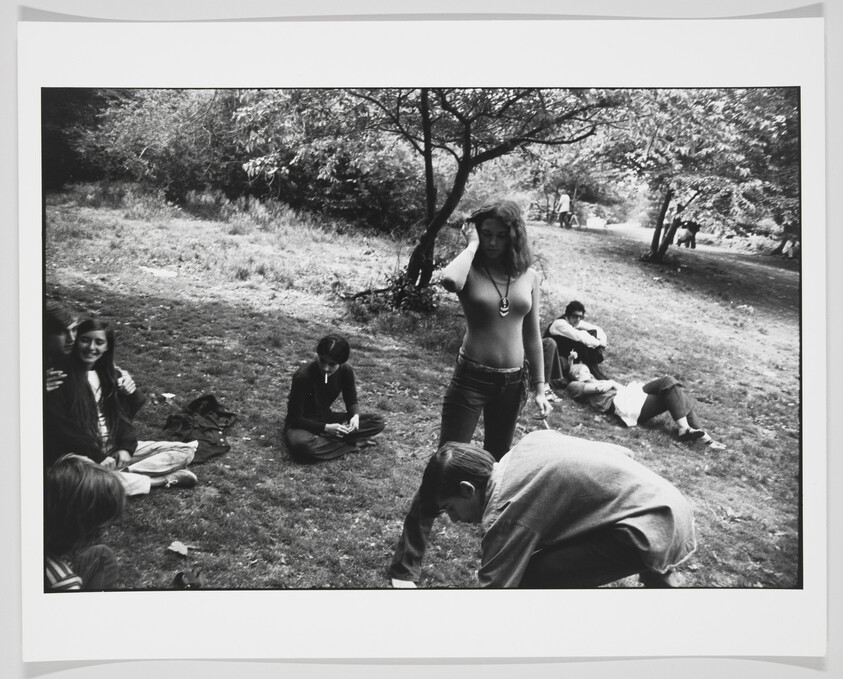 A black and white photograph capturing a casual outdoor scene with several people in a park-like setting. A woman stands in the center with her hand on her hip, wearing a necklace, while others are seated or lying on the grass around her, engaged in various activities such as reading or conversing. Trees and a pathway are visible in the background, suggesting a peaceful, leisurely environment.