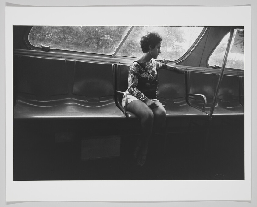 A black and white photograph of a woman sitting alone on a bus, gazing out of the window. She is dressed in a patterned top with short sleeves and a dark skirt, with her legs crossed. Her hair is styled in a short afro, and she wears large hoop earrings. The bus interior is empty except for her, highlighting a sense of solitude.