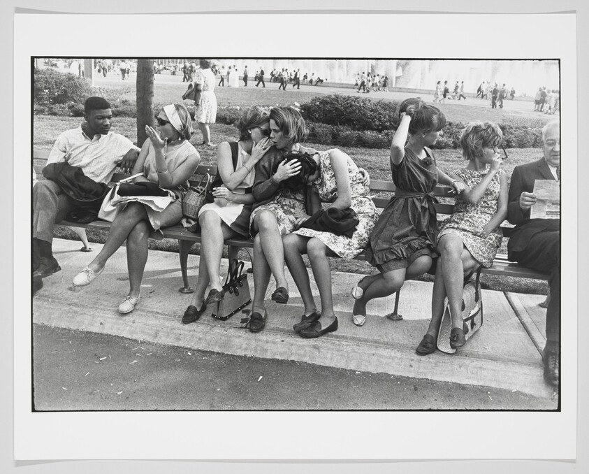 A black and white photograph of seven people sitting on a park bench. From left to right: a man and a woman engaged in conversation, two women whispering to each other, a woman fixing her hair, another woman lighting a cigarette, and an older man reading a newspaper. In the background, there are blurred figures of people walking in a park-like setting.