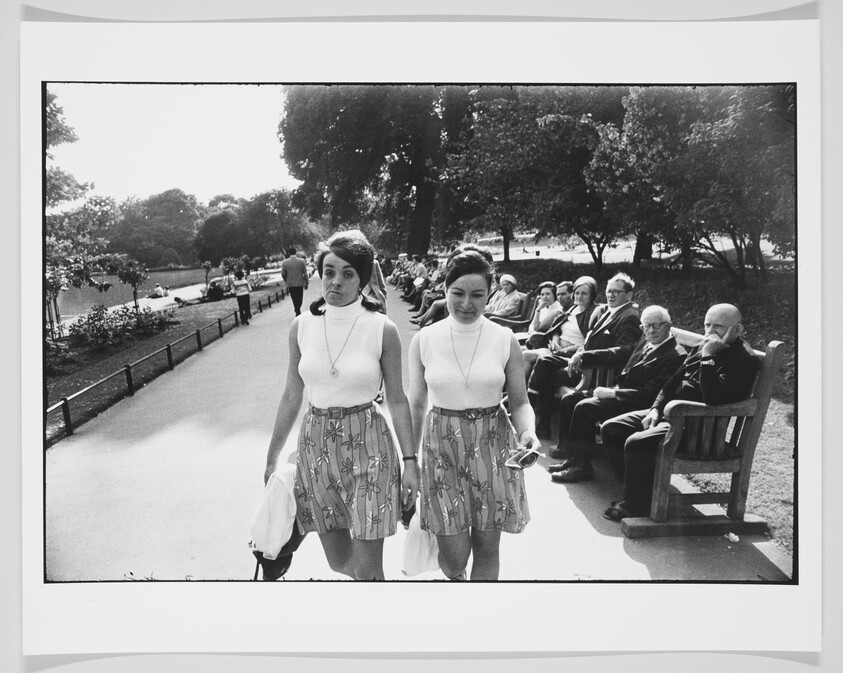 Black and white photograph of two young women walking side by side in a park, with one slightly ahead of the other. They are dressed in similar fashion with sleeveless tops and patterned skirts. In the background, several people, mostly elderly, are seated on benches, observing the passersby. Trees and a pond with ducks can be seen further in the distance.
