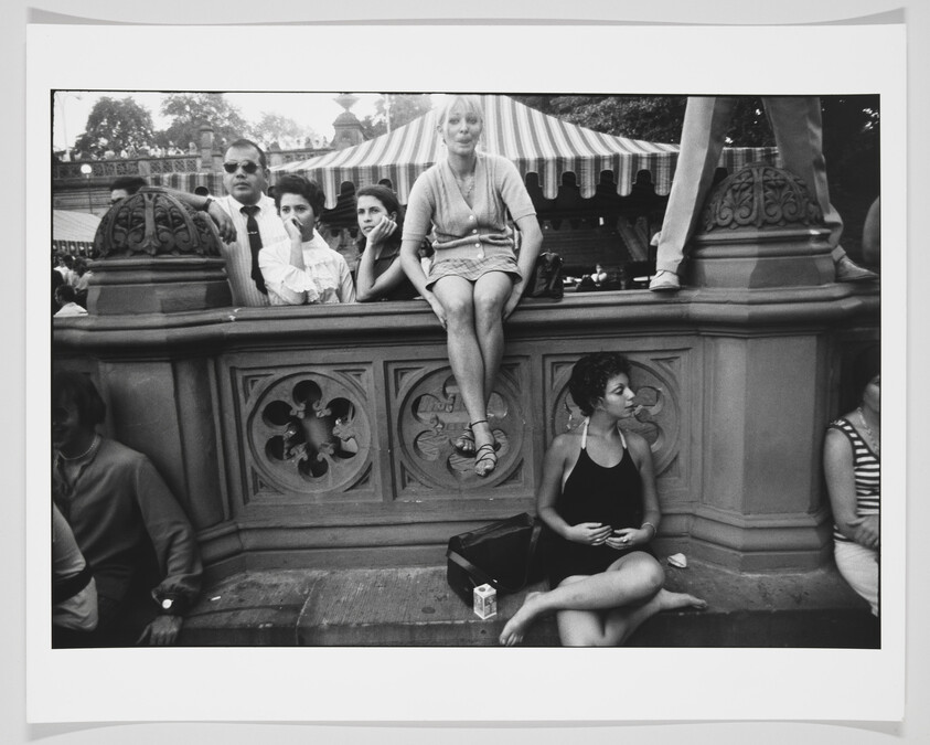 A black and white photograph capturing a diverse group of people sitting and standing around an ornate stone balustrade, with some individuals looking off into the distance and others engaged in various activities, such as eating or holding a camera. The setting appears to be a public outdoor area, possibly a park or near a street, with a striped tent in the background.