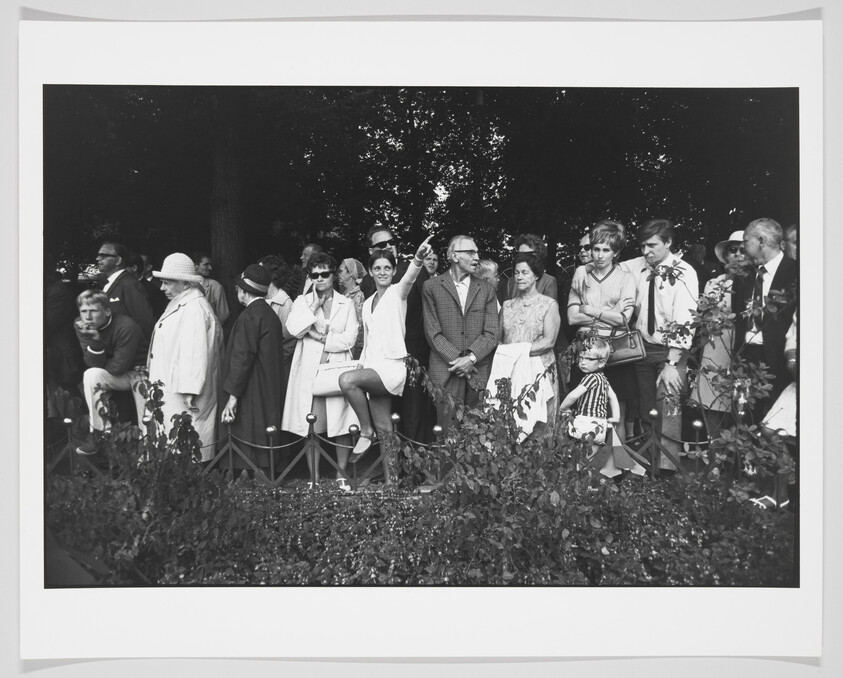 A black and white photograph of a diverse group of people standing behind a low fence, some seated on the fence, watching an event. The crowd appears engaged, with various expressions and styles of dress indicative of a past era.