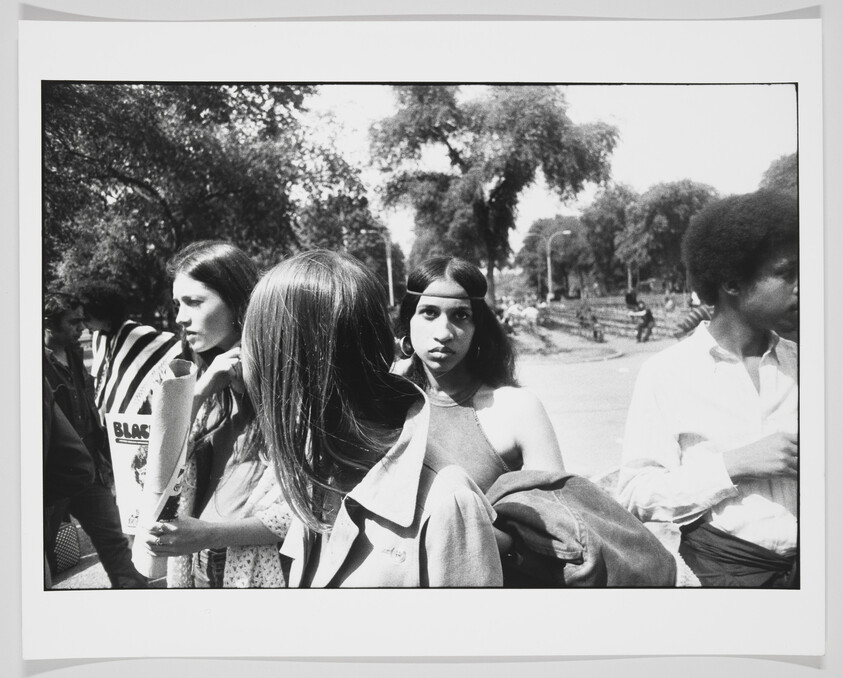 Black and white photograph capturing a candid moment among a group of young adults, possibly from the 1960s or 1970s. A woman in the center looks directly at the camera with a headband in her hair, while others are engaged in conversation around her. A man on the right side is partially out of frame, and the background shows a park-like setting with trees and a path.