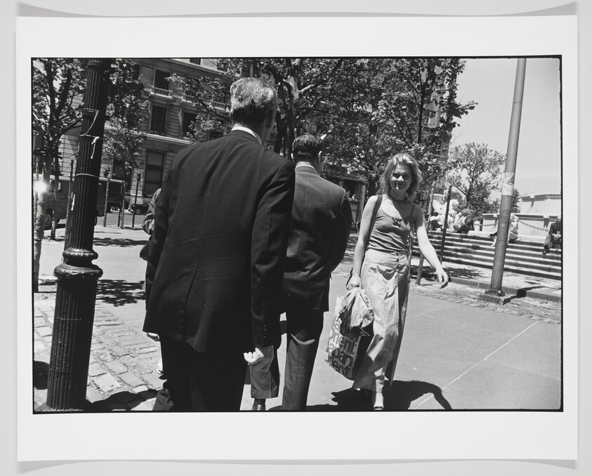 A black and white photograph capturing a moment on a sunny city street where two men in business suits are seen from behind, walking to the left, and a woman with sunglasses and a shoulder bag is walking towards the right, looking directly at the camera. The background features urban trees, a street lamp, and the edge of a park with steps and a railing.