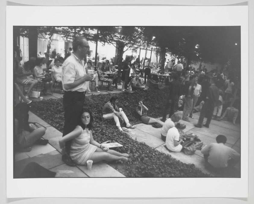 A black and white photograph capturing a casual outdoor gathering with various people sitting and standing, some on steps and others on grass. A man in the foreground stands holding a drink, while others in the background engage in conversation, relaxation, or pass by. The scene suggests a social event in a park-like setting with trees and foliage around.