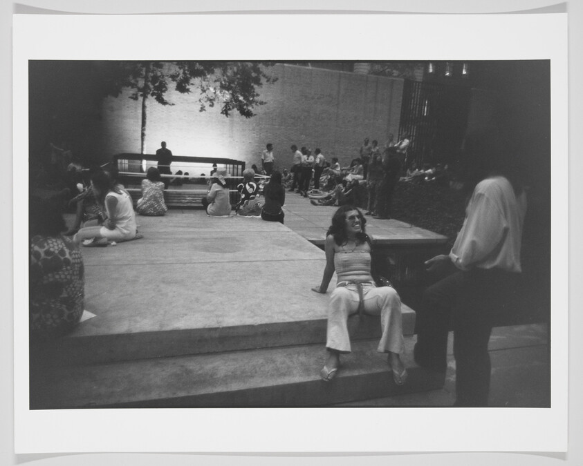 A black and white photograph capturing a casual outdoor scene where various people are sitting and lounging on steps and benches. In the foreground, a woman in light-colored clothing is seated on a large step, smiling and engaging with someone standing in front of her, who is blurred in motion. The background shows a mix of individuals and small groups in a relaxed social setting, with trees and a brick wall providing a backdrop.