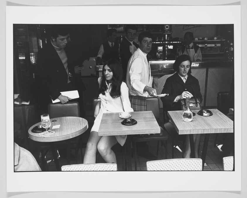 Black and white photograph of a vintage café scene with two women seated at separate tables. The woman on the left is dressed in a white outfit and is looking to the side, with a glass of beer and an ashtray on her table. The woman on the right is wearing a dark blazer and skirt, taking notes, with a carafe and cup on her table. In the background, a man in a white shirt is serving, and another man in a dark jacket is observing the scene.