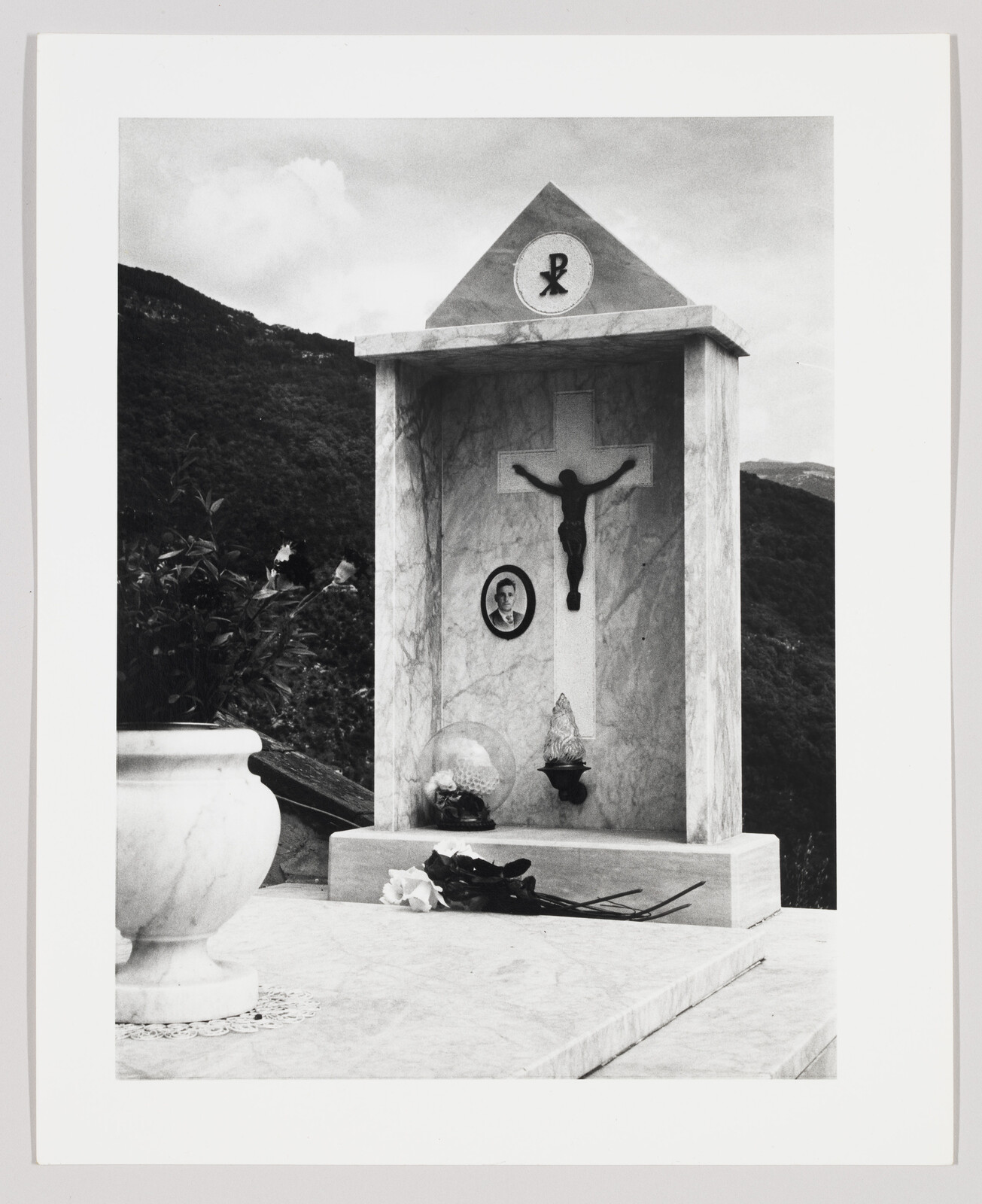 A marble roadside shrine with a crucifix, a small portrait, and wilted flowers laid on the base.