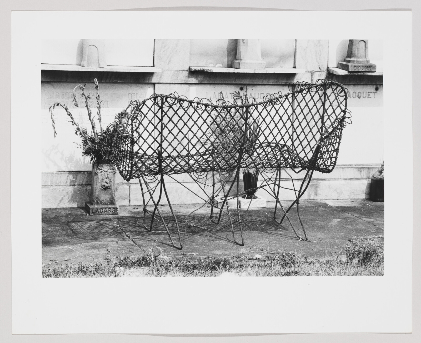 A woven metal bench with plants and a vase set against a stone wall.