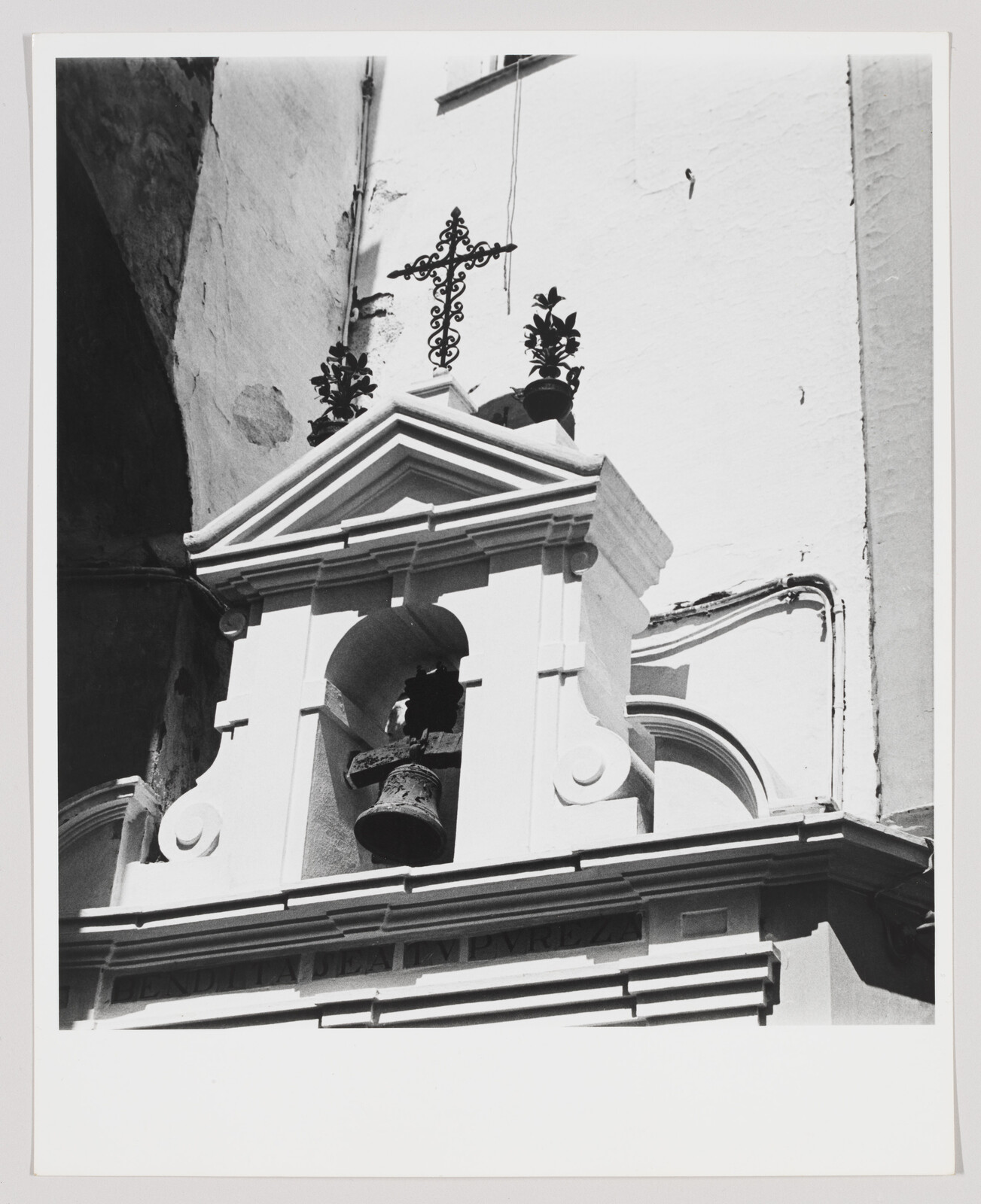 Small bell hangs in a white church niche beneath an ornate metal cross and potted decorations.
