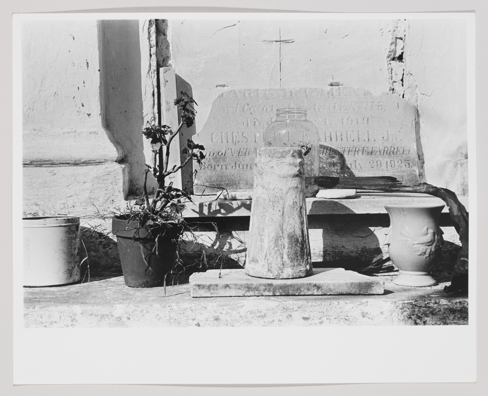 Weathered grave marker with mason jar, potted plant, and decorative vase arranged on a ledge.