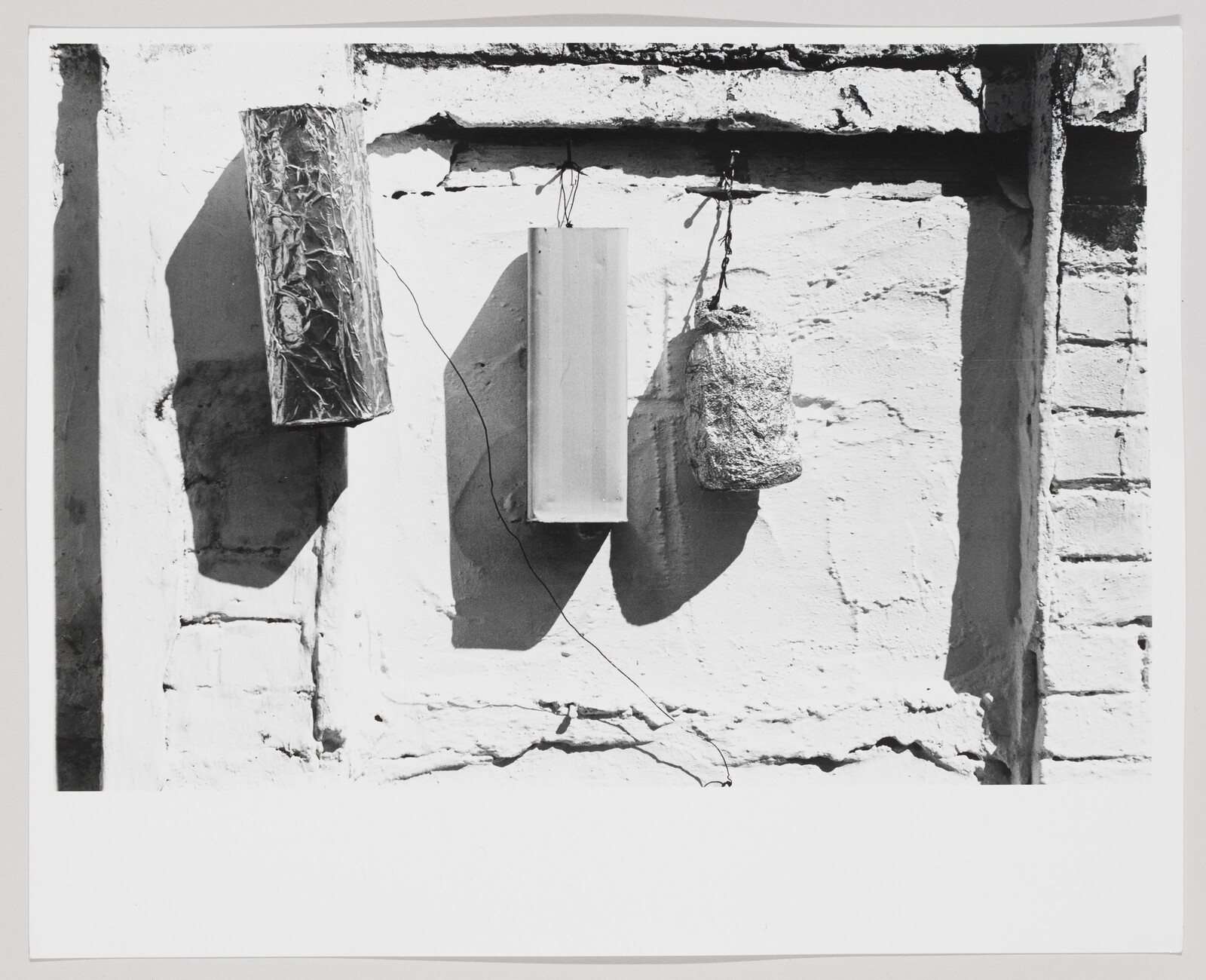 Three objects hang from a wall, casting distinct shadows on the sunlit plaster.