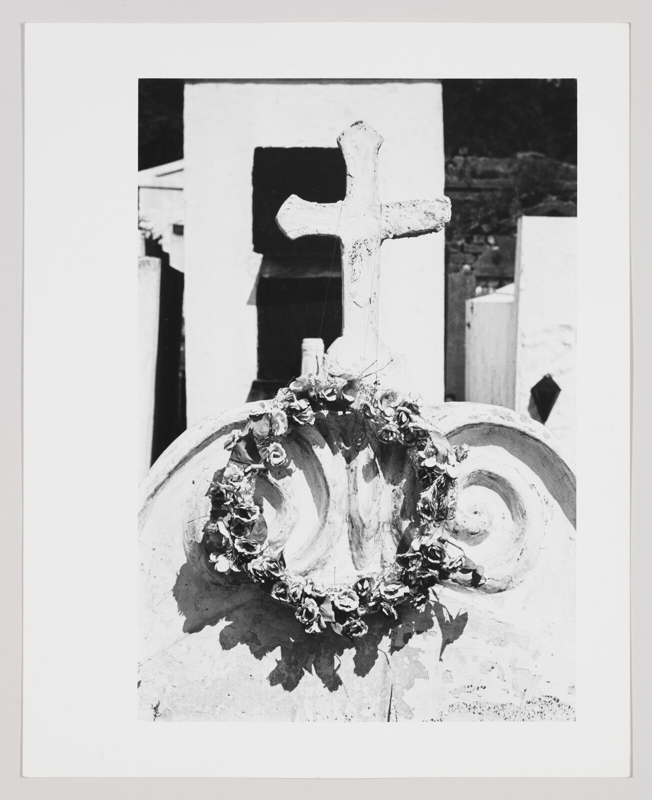 A weathered stone grave marker topped by a cross and draped with a wreath of flowers.