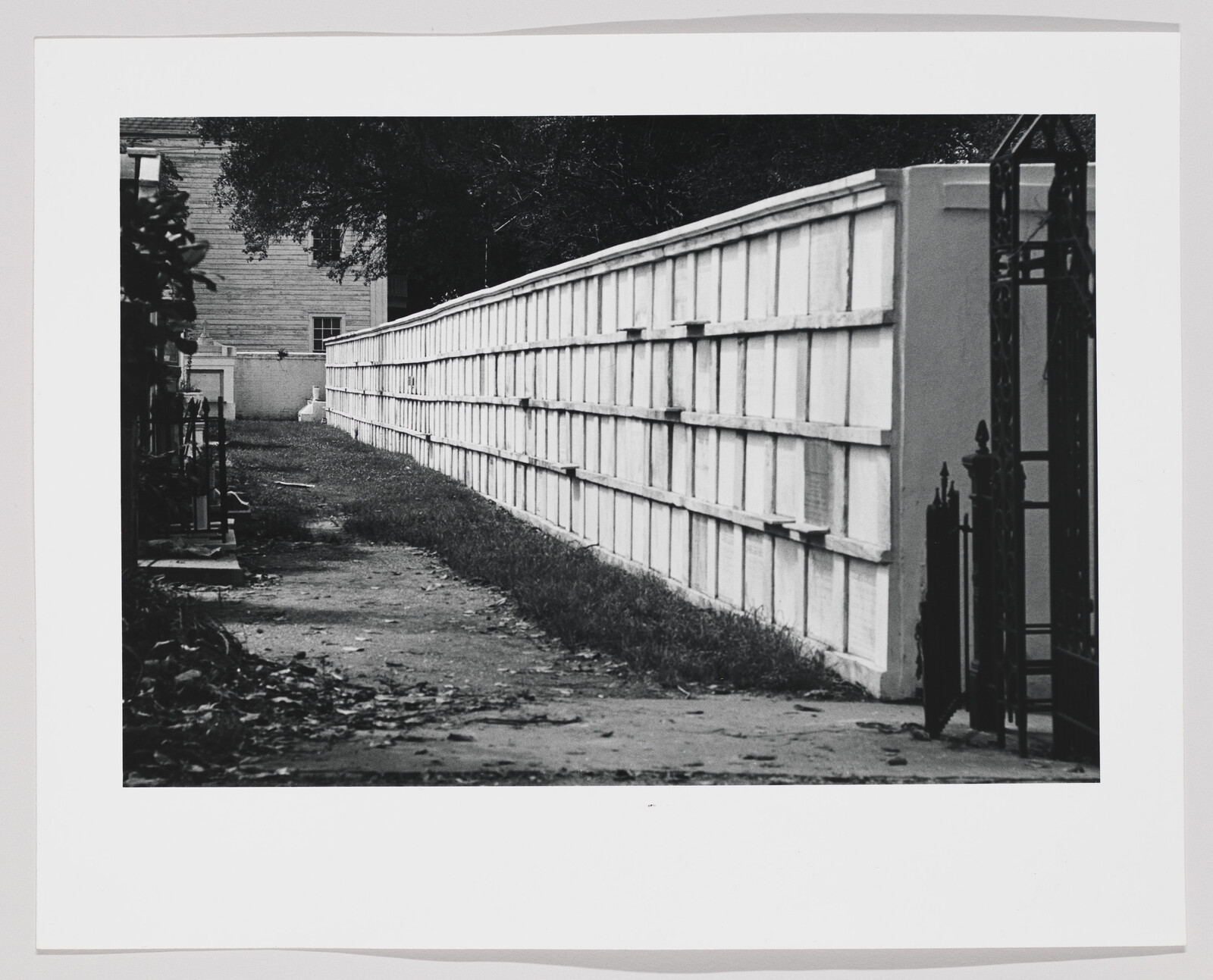 A long white columbarium wall lines a narrow grassy walkway beside an open iron gate.