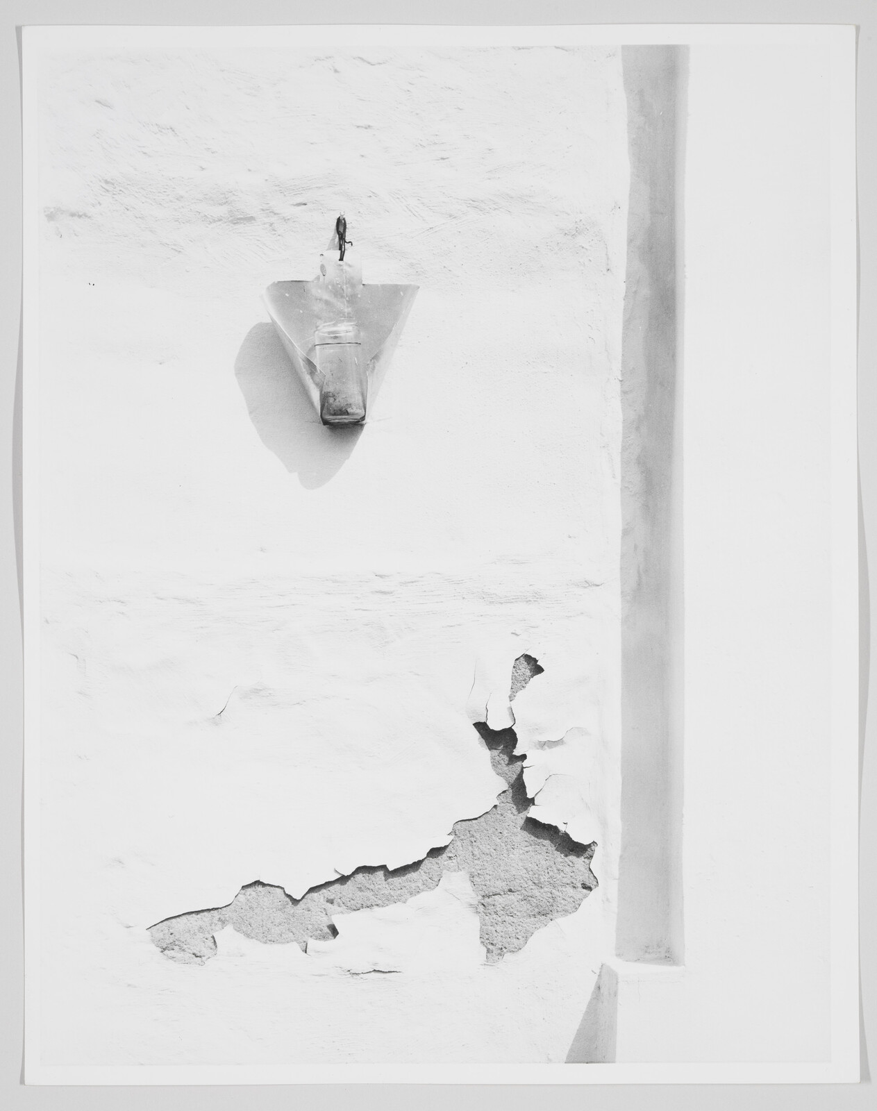 A small outdoor lamp casts a shadow above peeling plaster and cracked paint on a white wall.