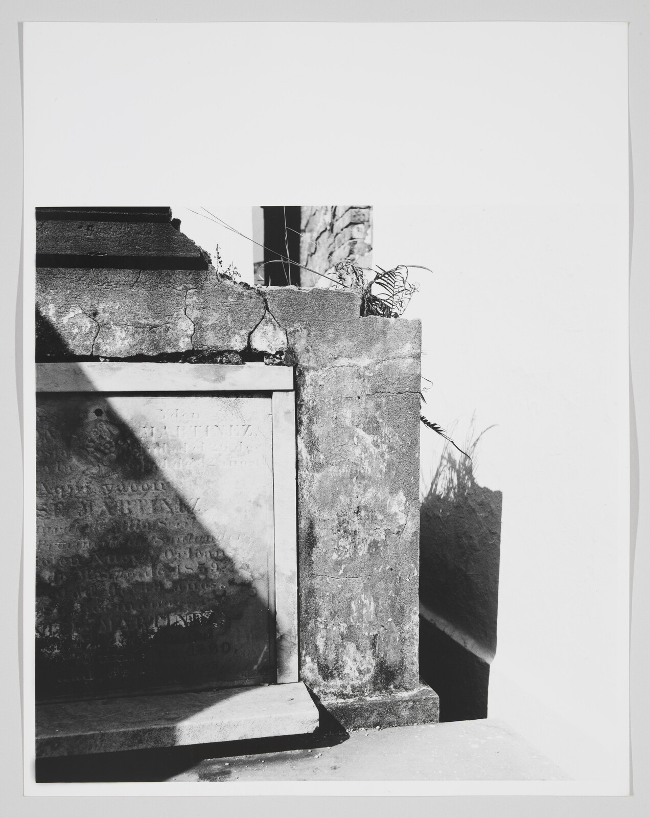 Weathered stone tomb with cracked pillar, sunlight casting diagonal shadows and small plants.