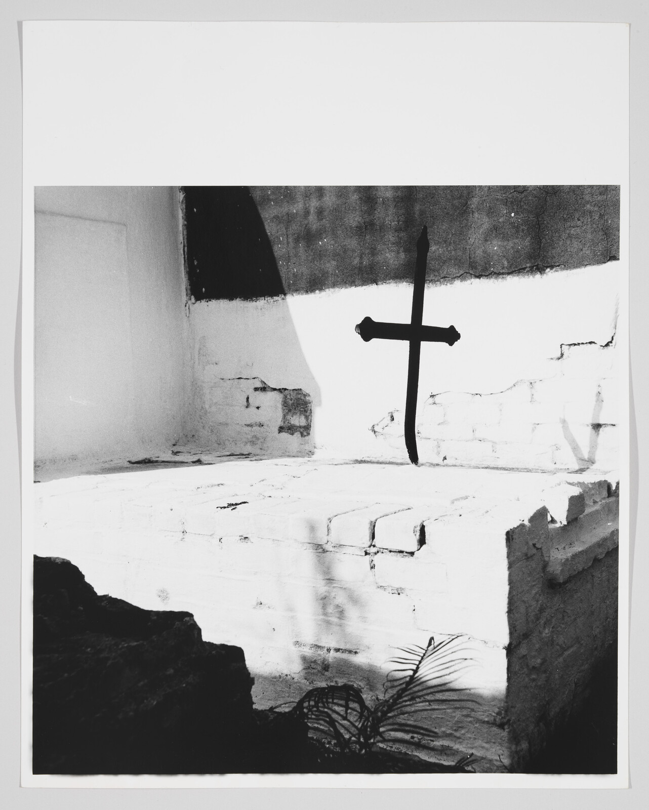 A dark iron cross mounted on a sunlit white brick wall casting sharp shadows.