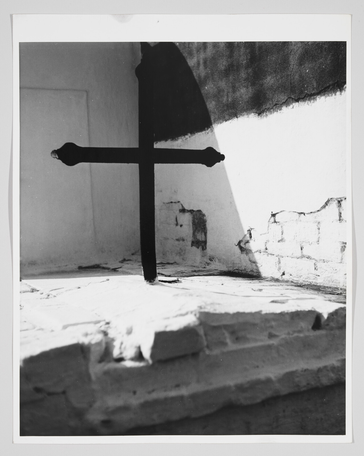 A dark metal cross stands on a sunlit plaster ledge casting a sharp shadow on the wall.