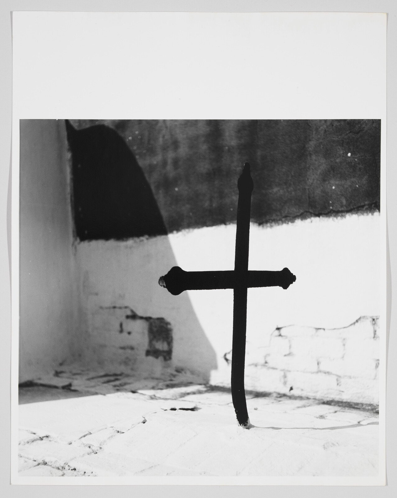 A simple black metal cross stands upright casting a sharp shadow on a white wall and floor.