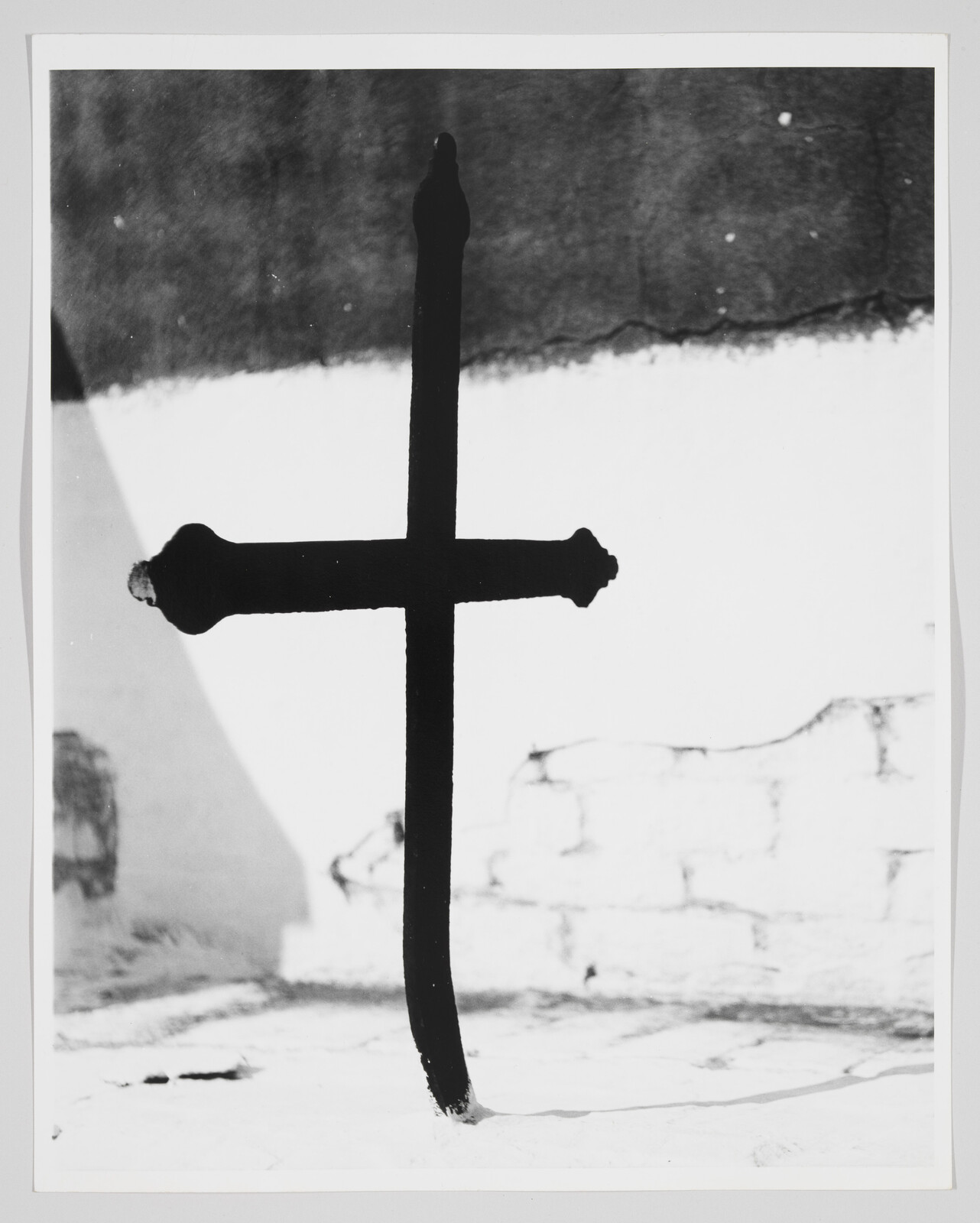 A tall black iron cross planted in bright white ground casting a shadow on the wall.