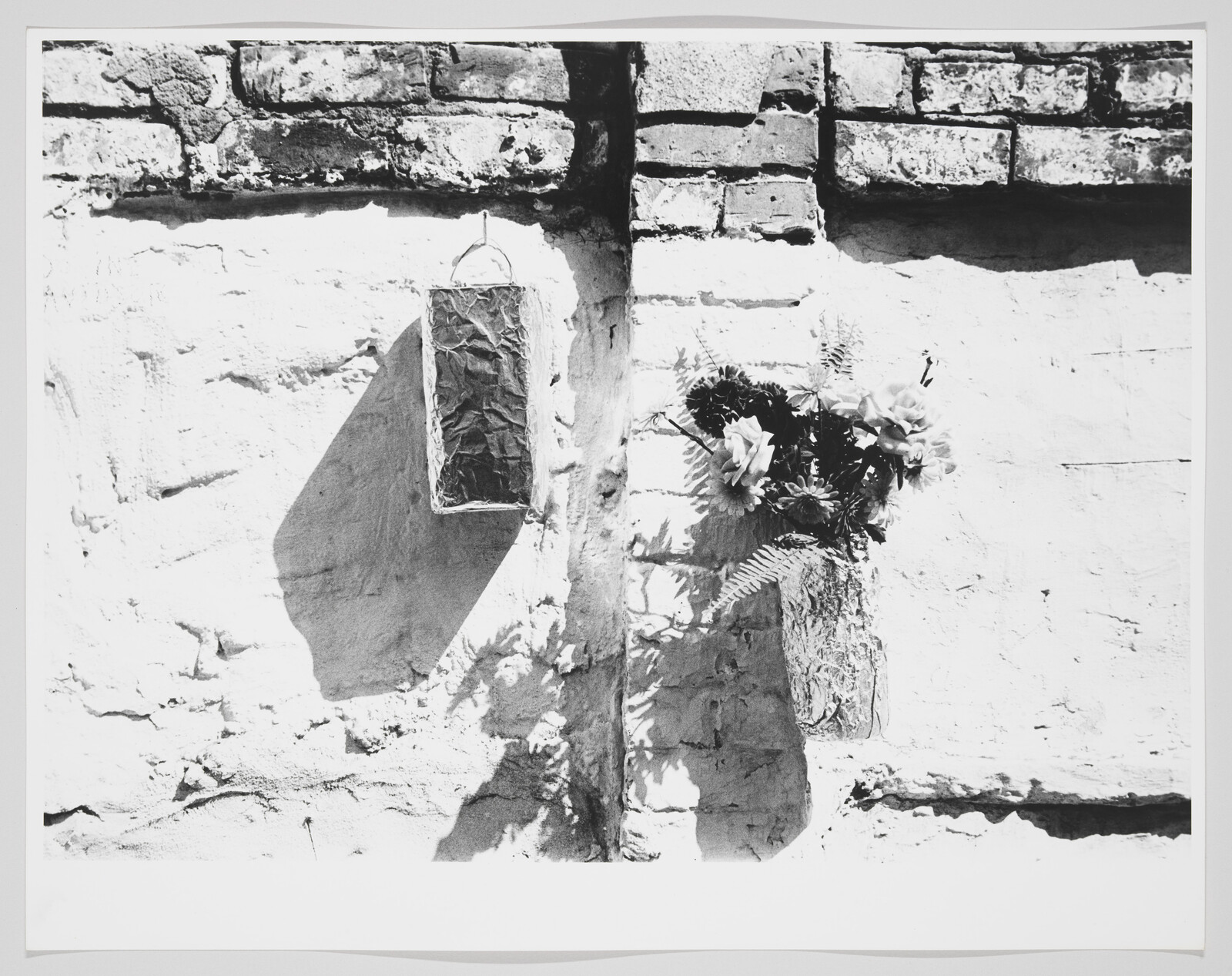 A weathered brick wall with a hanging rectangular planter casting a shadow and a vase of flowers.