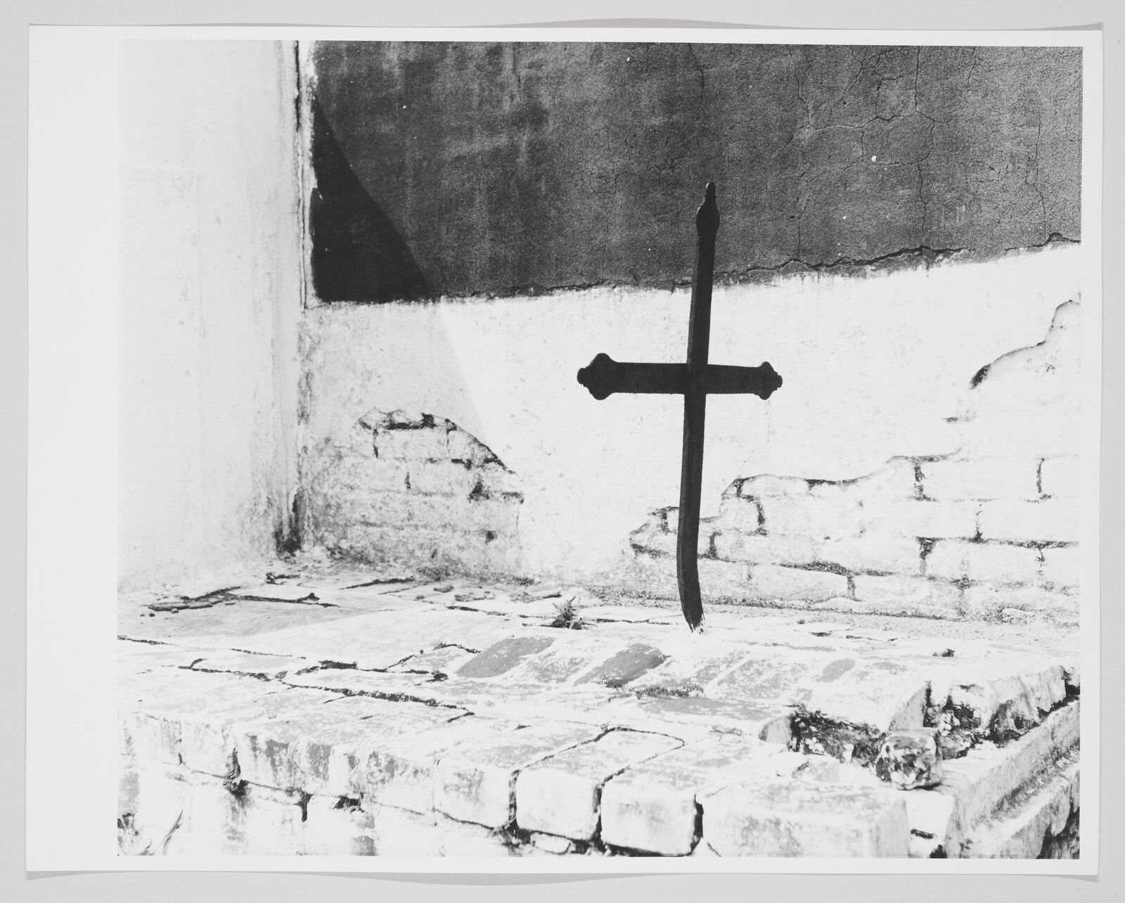 A black iron cross mounted on a cracked white wall above an old brick platform.