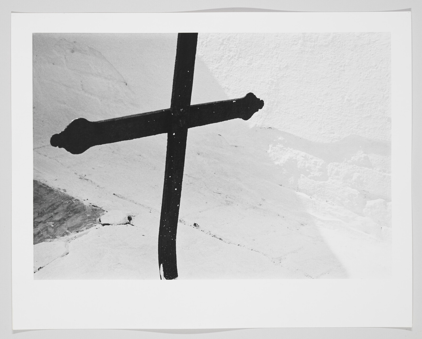 A dark iron cross leans against a white textured wall with visible cracks and shadows.