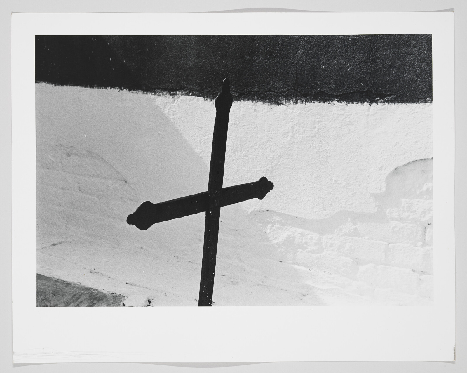 A black metal cross stands tilted against a textured white wall with a dark upper band.