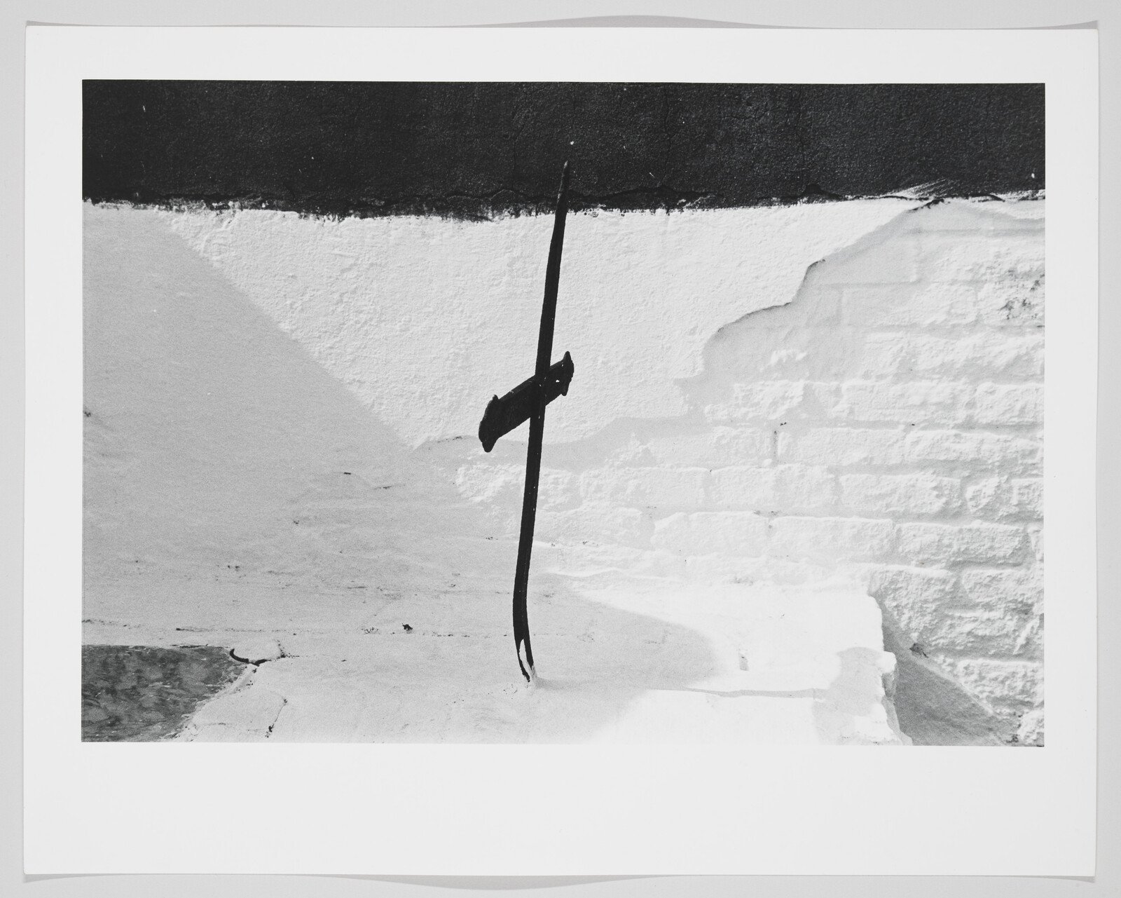 A thin black metal stake casts a shadow against a white textured brick wall.